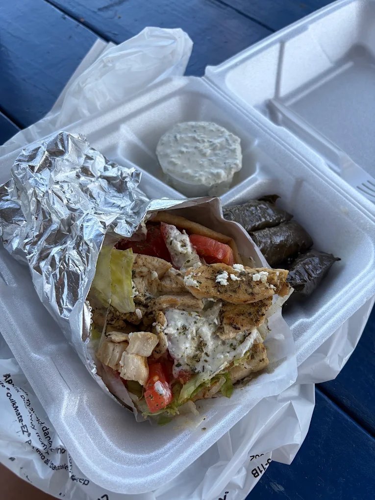 A chicken gyro in foil in a takeout container — grilled chicken, feta, tomato and lettuce — beside three dolmades and a cup of tzatziki
