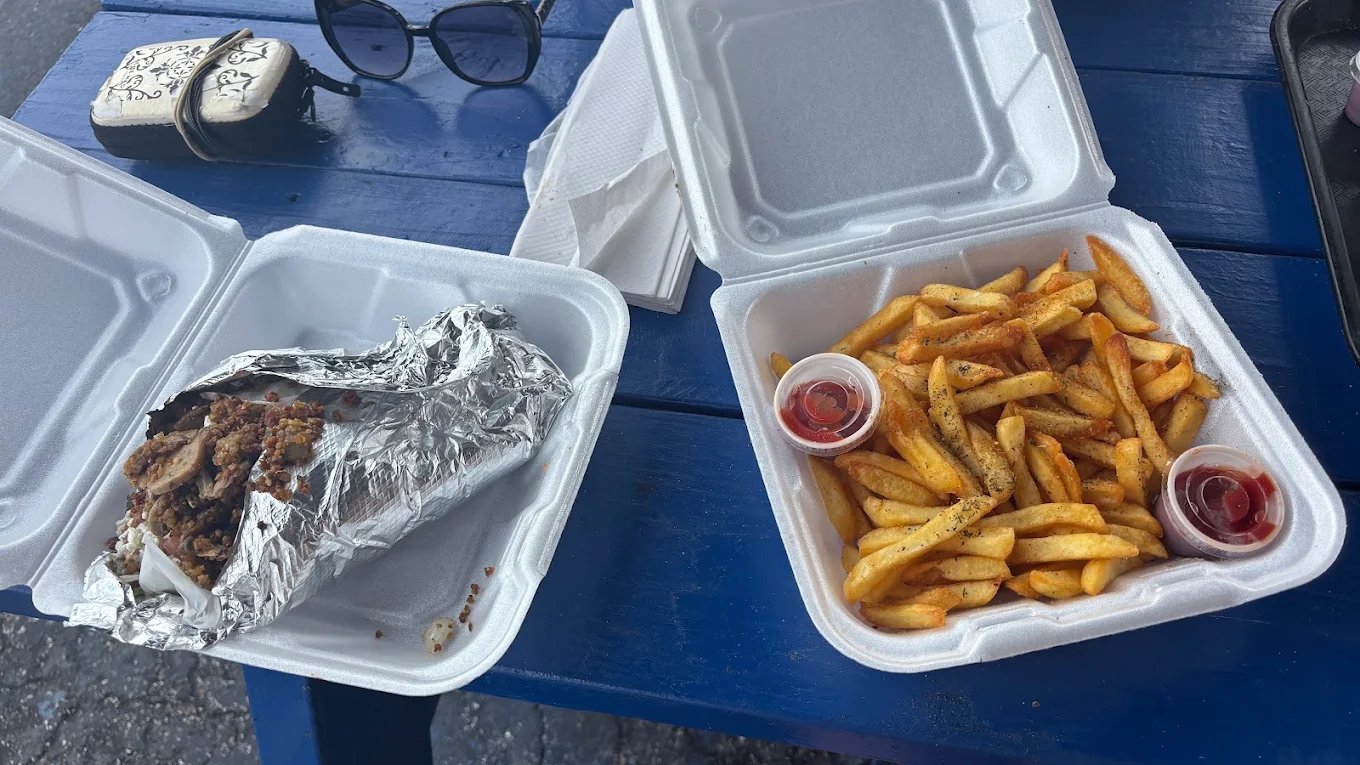 Two open takeout containers on a blue picnic table — a foil-wrapped gyro in one, a large portion of seasoned Greek fries with two cups of ketchup in the other