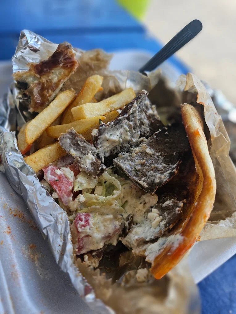 An opened foil tray holding sliced gyro meat, french fries, lettuce, tomato and tzatziki, with a plastic fork resting in it, on a blue picnic table