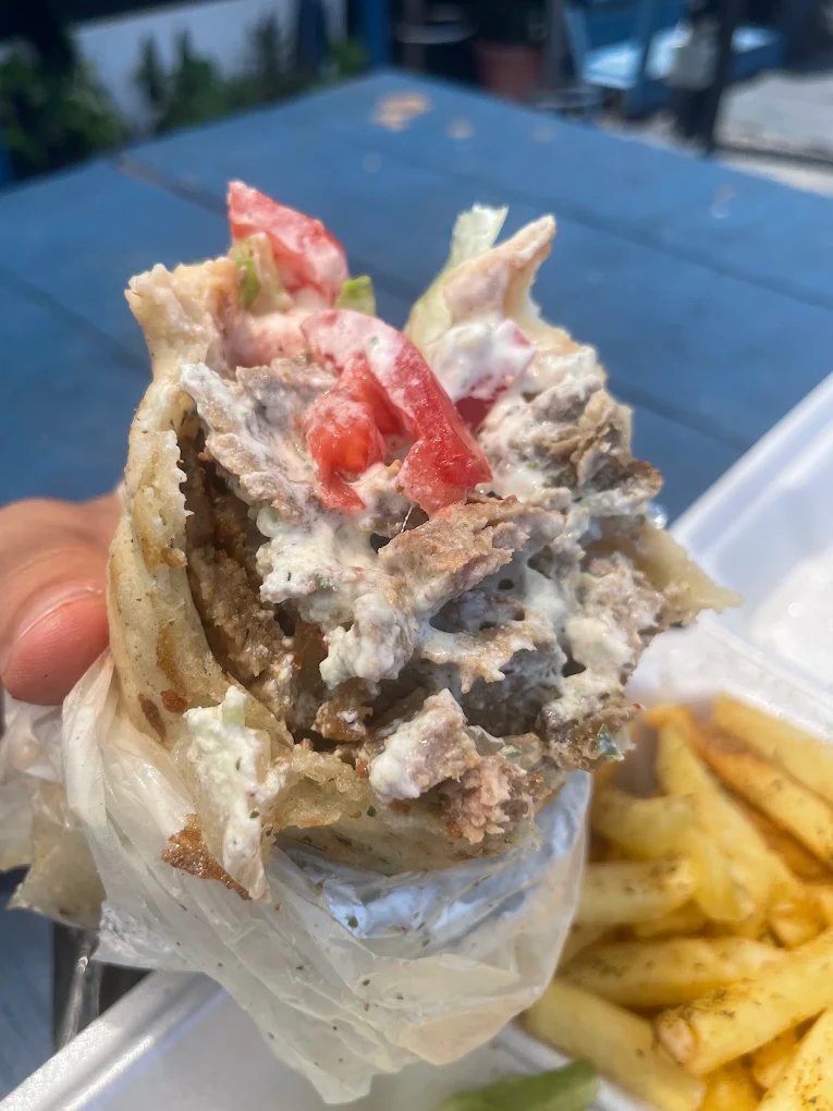 A hand holding a paper-wrapped gyro, open at the top to show sliced gyro meat, tzatziki, chopped tomato and lettuce, with a container of fries behind it