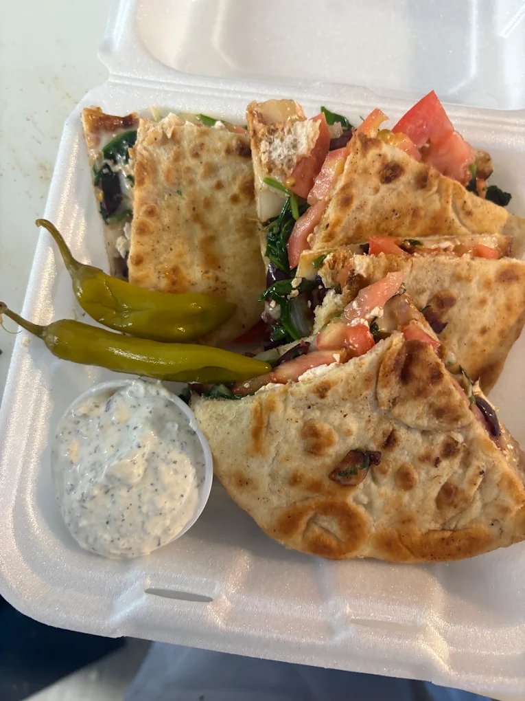 Wedges of spinach and feta pie in a takeout container with chopped tomato, two pepperoncini peppers and a cup of tzatziki